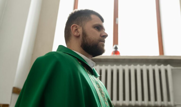 A bearded priest in a green liturgical garment stands indoors, reflecting religious significance.