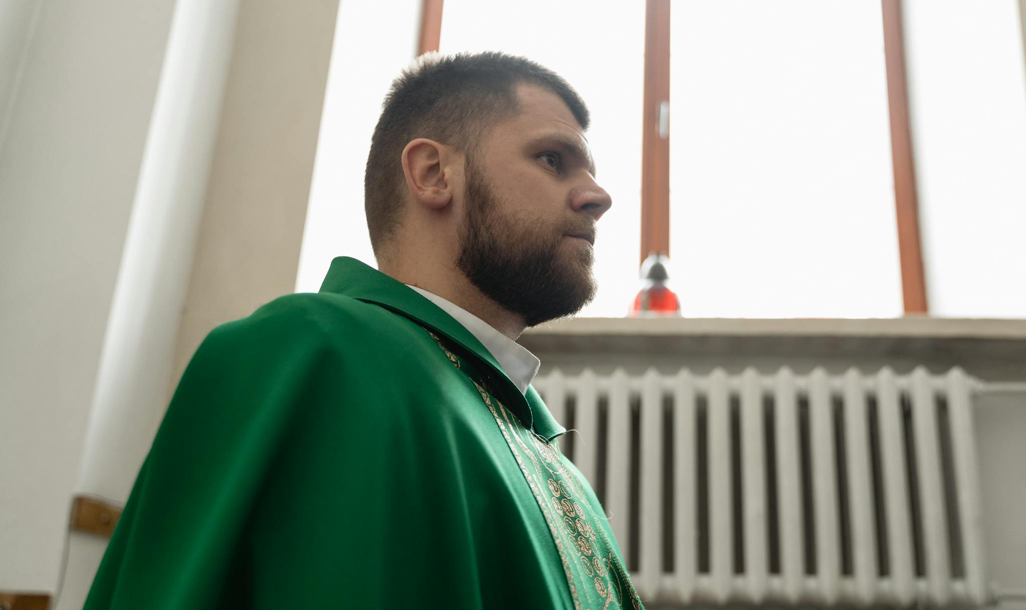 A bearded priest in a green liturgical garment stands indoors, reflecting religious significance.